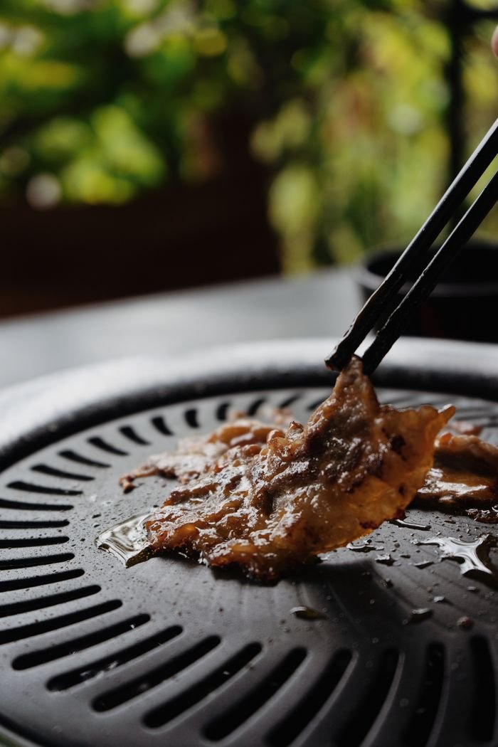 Close-up of grilled meat held by chopsticks over a barbecue grill outdoors.